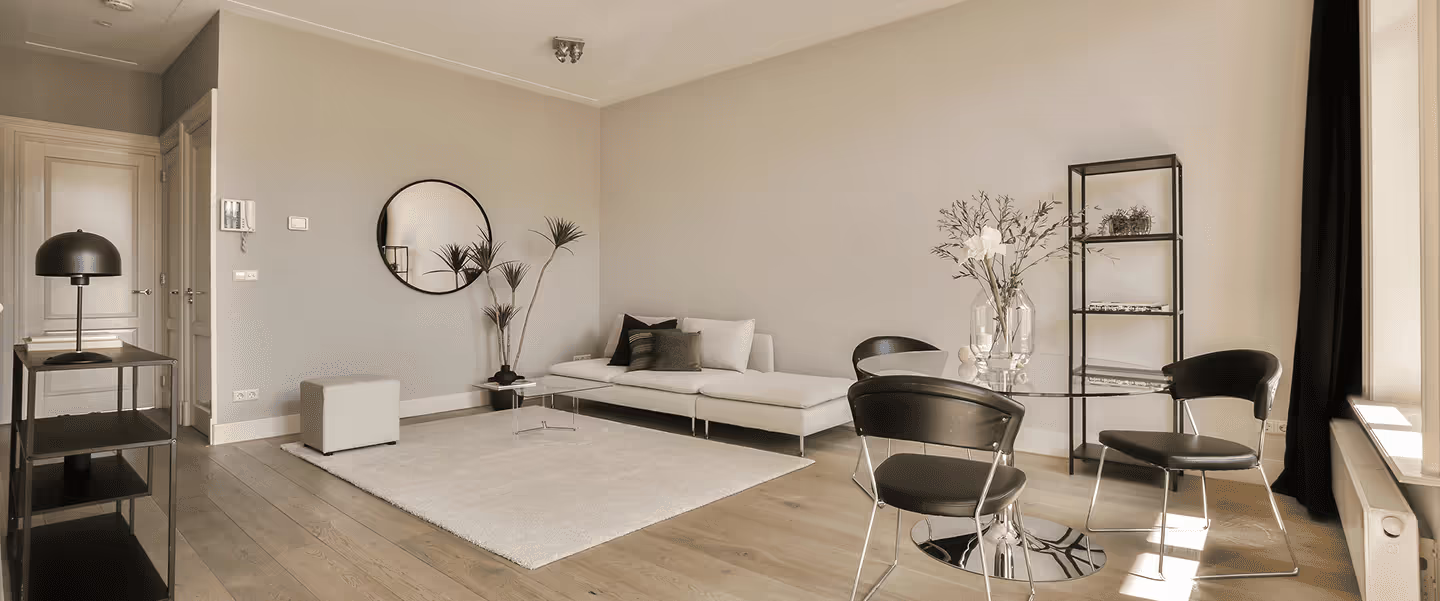 Modernes Wohnzimmer mit weißem Sofa, rundem Glasesszimmertisch, schwarzen Stühlen, einem Regal und Pflanzen vor heller Wand.