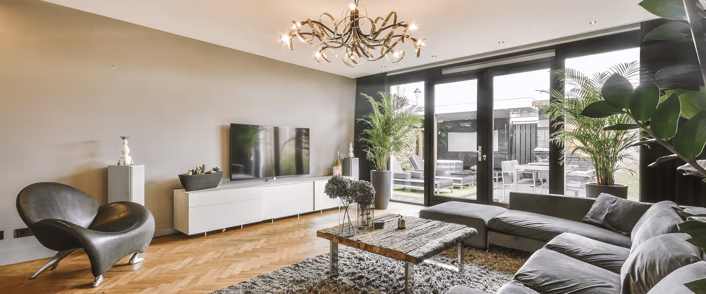 Modernes Wohnzimmer mit großem grauem Ecksofa, Holztisch, dunkelgrauem Sessel, großem Fernseher und bodentiefen Fenstern mit Blick auf eine Terrasse.