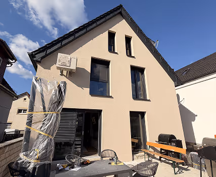 Haus mit drei großen Fenstern und einer Terrasse mit Gartenmöbeln unter blauem Himmel.