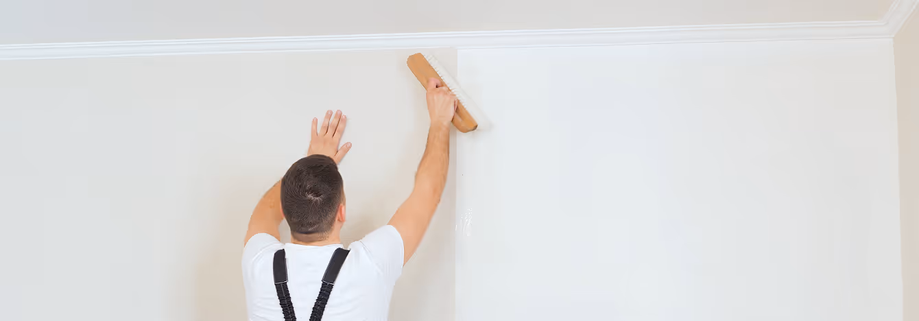 Ein Mann in weißem T-Shirt und schwarzen Hosenträgern glättet eine weiße Wand mit einem Werkzeug.