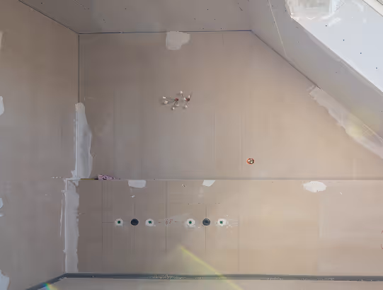 Unfertiger Innenraum mit trockenbauwänden, sichtbaren Steckdosen und einem Dachfenster, das Tageslicht hereinlässt.