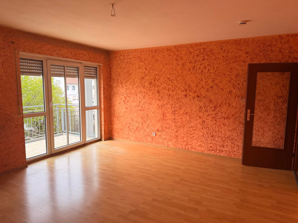 Leeres Wohnzimmer vor der Renovierung mit orangefarbenen, grob strukturierten Wänden und Balkontür